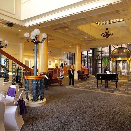 Stamford Grand Adelaide Hotel Glenelg Interior foto The photo shows a luxurious interior of a hotel or a restaurant lobby. There is an elegant staircase with ornate railings leading to an upper level. The space features high ceilings with decorative chandeliers and ample natural light coming through l