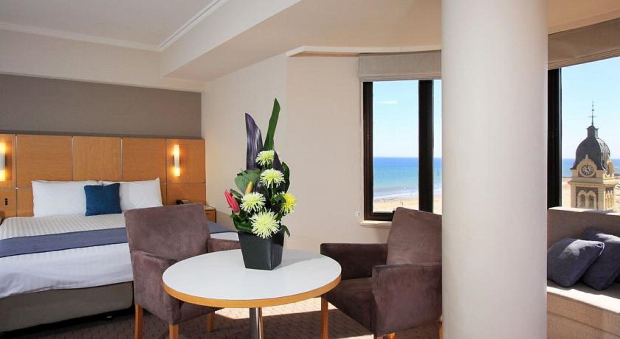 Stamford Grand Adelaide Hotel Glenelg Exterior foto The photo shows a cozy hotel room with a comfortable seating arrangement. There are two brown chairs positioned around a round table, which features a decorative flower arrangement in a black vase. The background includes a window that offers a view 