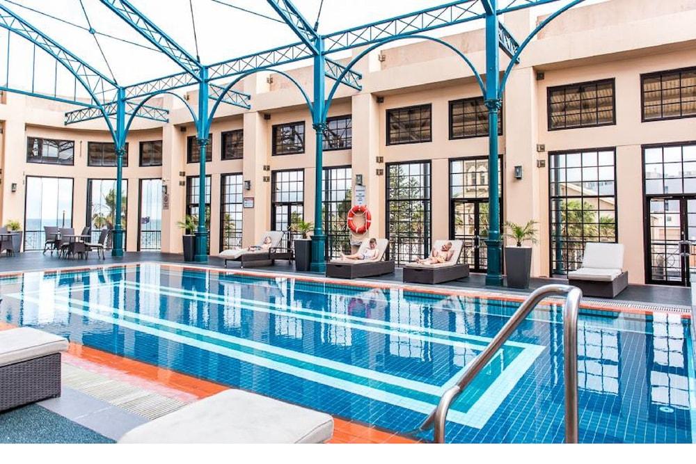 Stamford Grand Adelaide Hotel Glenelg Exterior foto The photo shows an indoor swimming pool area characterized by a modern and elegant design. The pool features blue tiles with two white stripes running along its length. Surrounding the pool, there are several lounge chairs for relaxation. The walls h