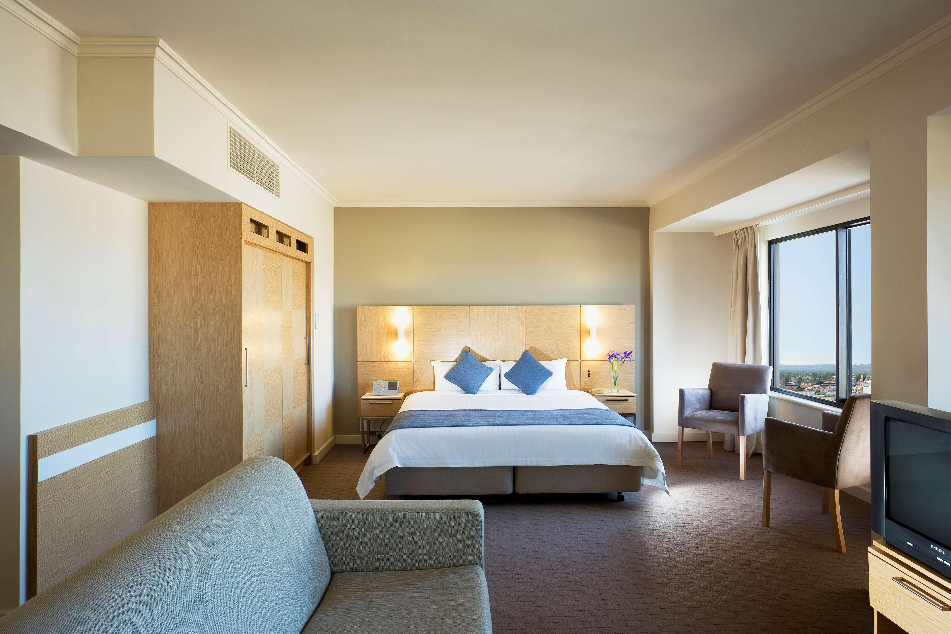 Stamford Grand Adelaide Hotel Glenelg Quarto foto The photo shows a modern hotel room featuring a large bed with two blue decorative pillows. There are bedside lamps on both sides of the headboard. To the left, there's a wooden wardrobe and a small table with a vase and a few amenities. On the right