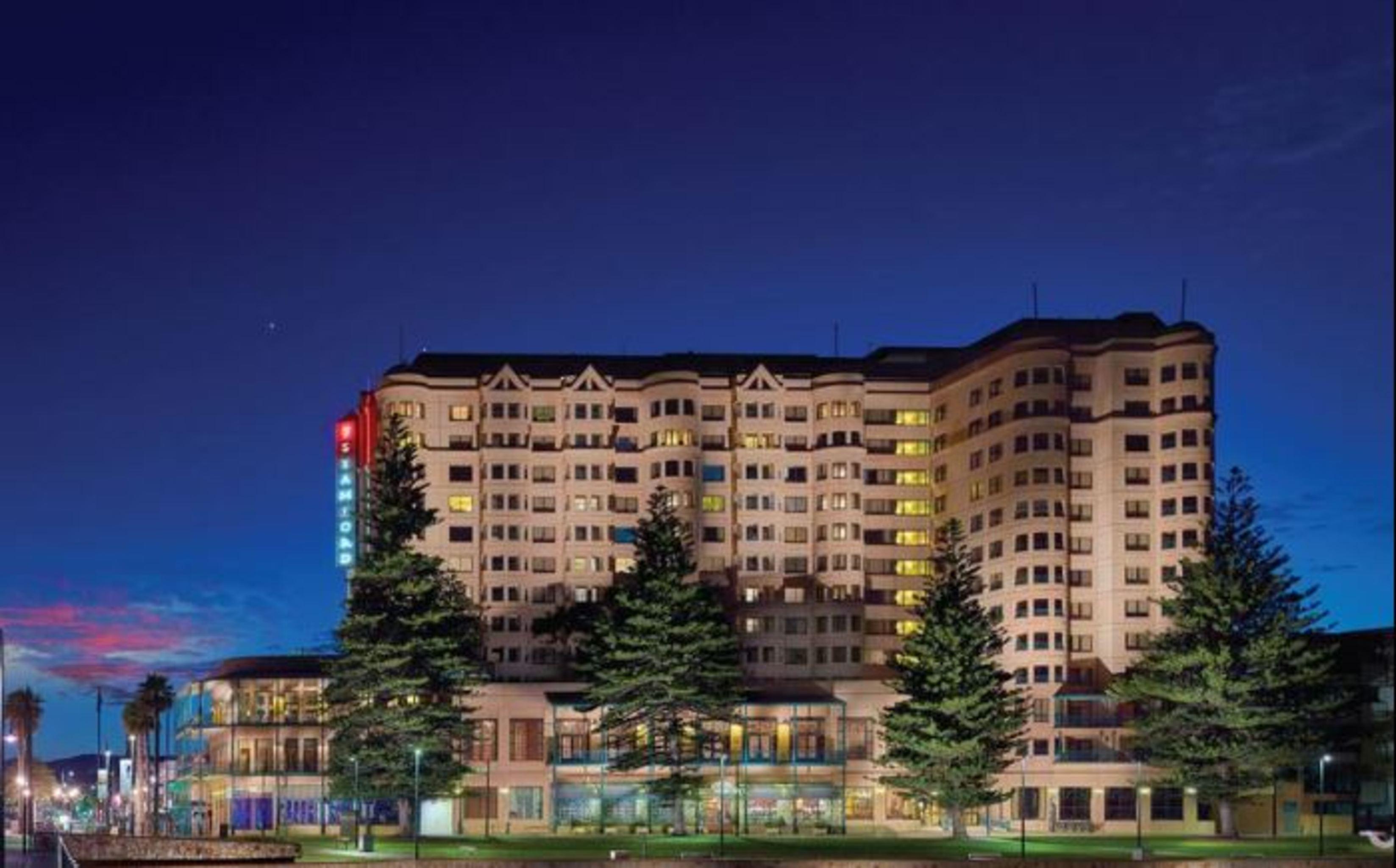 Stamford Grand Adelaide Hotel Glenelg Exterior foto The photo displays a multi-story hotel building at dusk, illuminated by various lights. The architecture features a mix of modern and traditional design elements, with numerous windows showcasing a warm glow. Surrounding the hotel are several tall tr