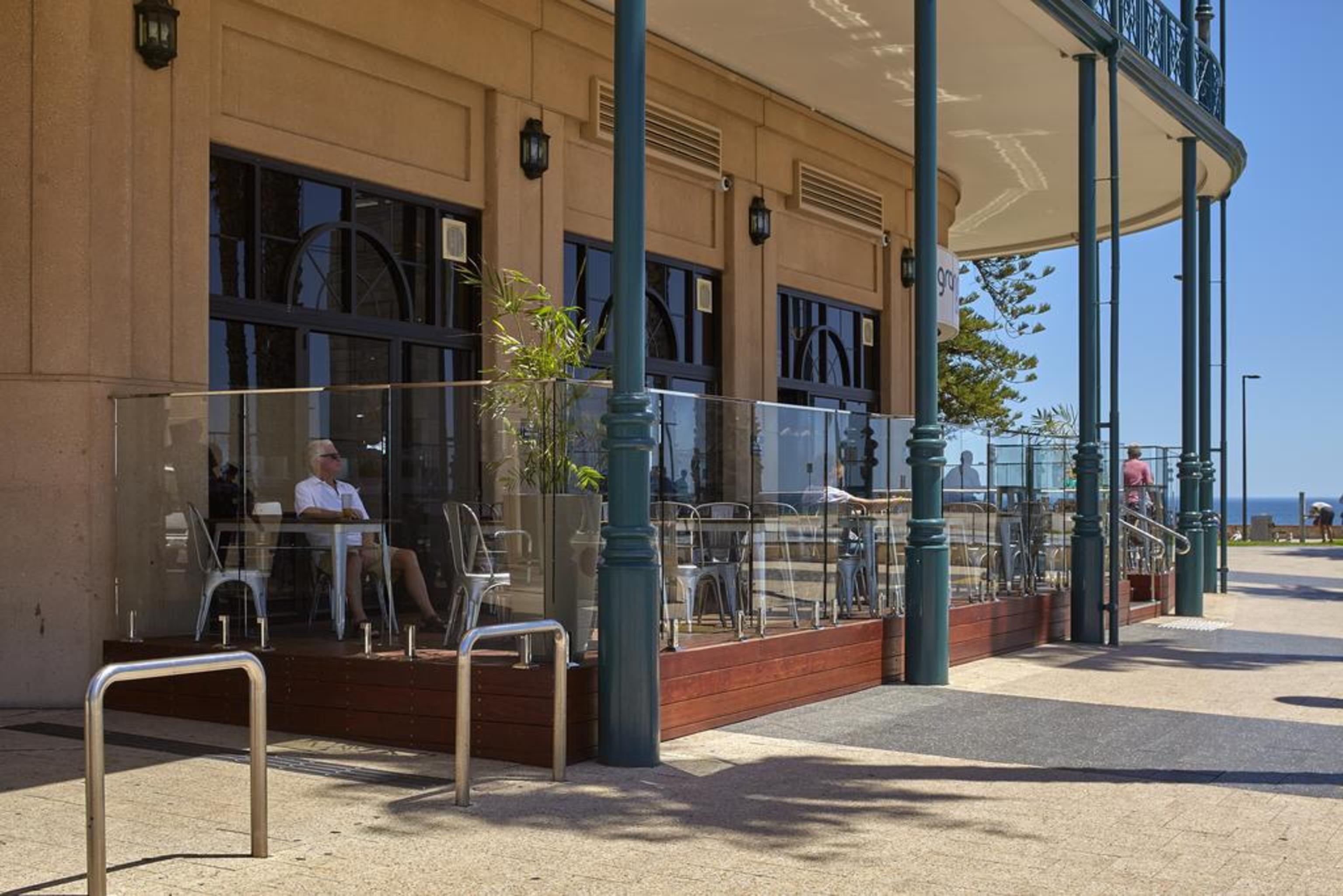 Stamford Grand Adelaide Hotel Glenelg Exterior foto The photo shows the exterior of a cafe or restaurant with a large outdoor seating area. There are glass barriers around the seating, providing a view of the outside while maintaining a sense of enclosure. The building has a modern architectural style