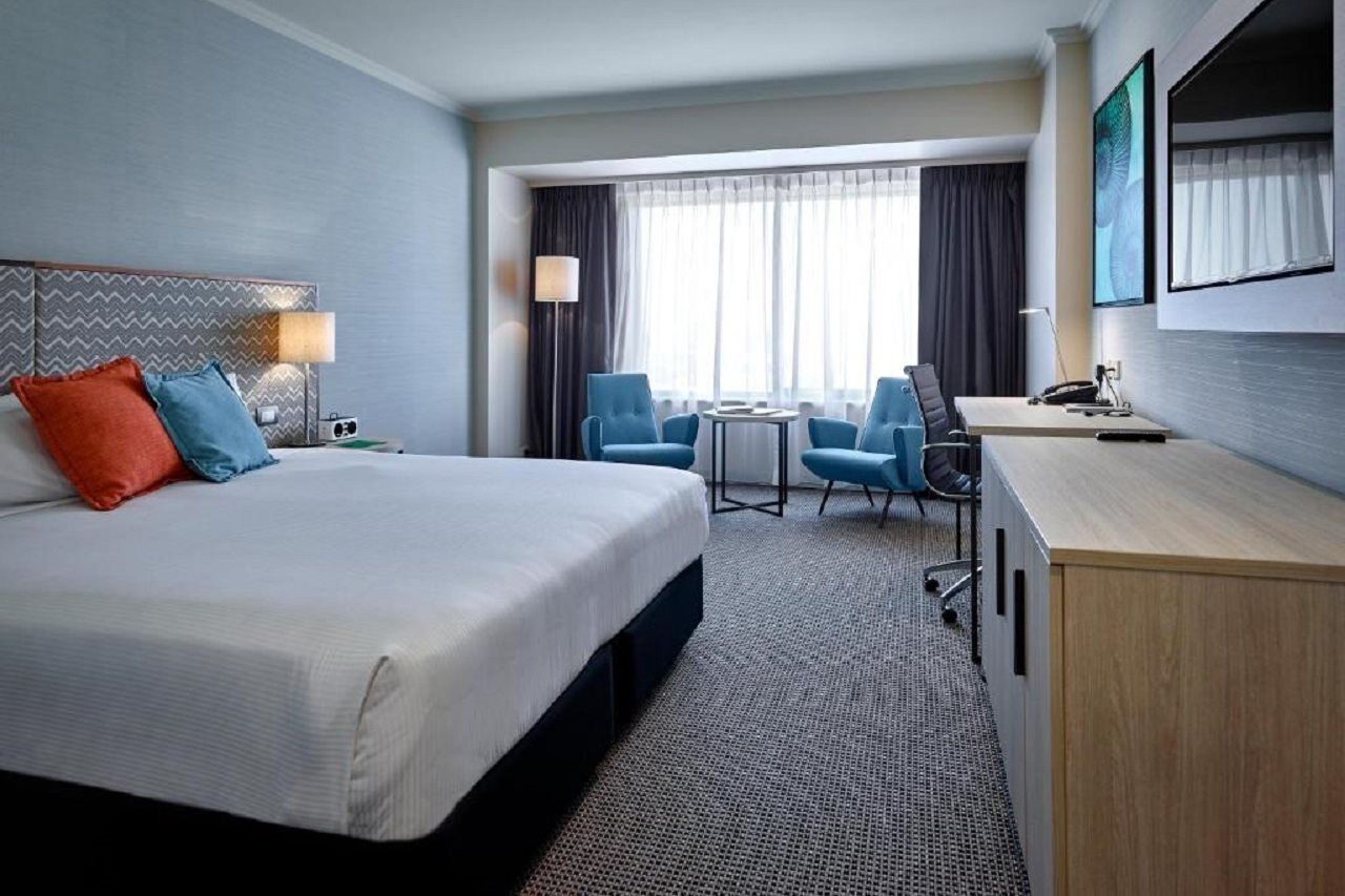 Stamford Grand Adelaide Hotel Glenelg Exterior foto The photo shows a modern hotel room with a clean and minimalist design. In the center, there is a large, neatly made bed with white linens and two decorative pillows, one orange and one blue. To the right, there is a small wooden cabinet with a telev